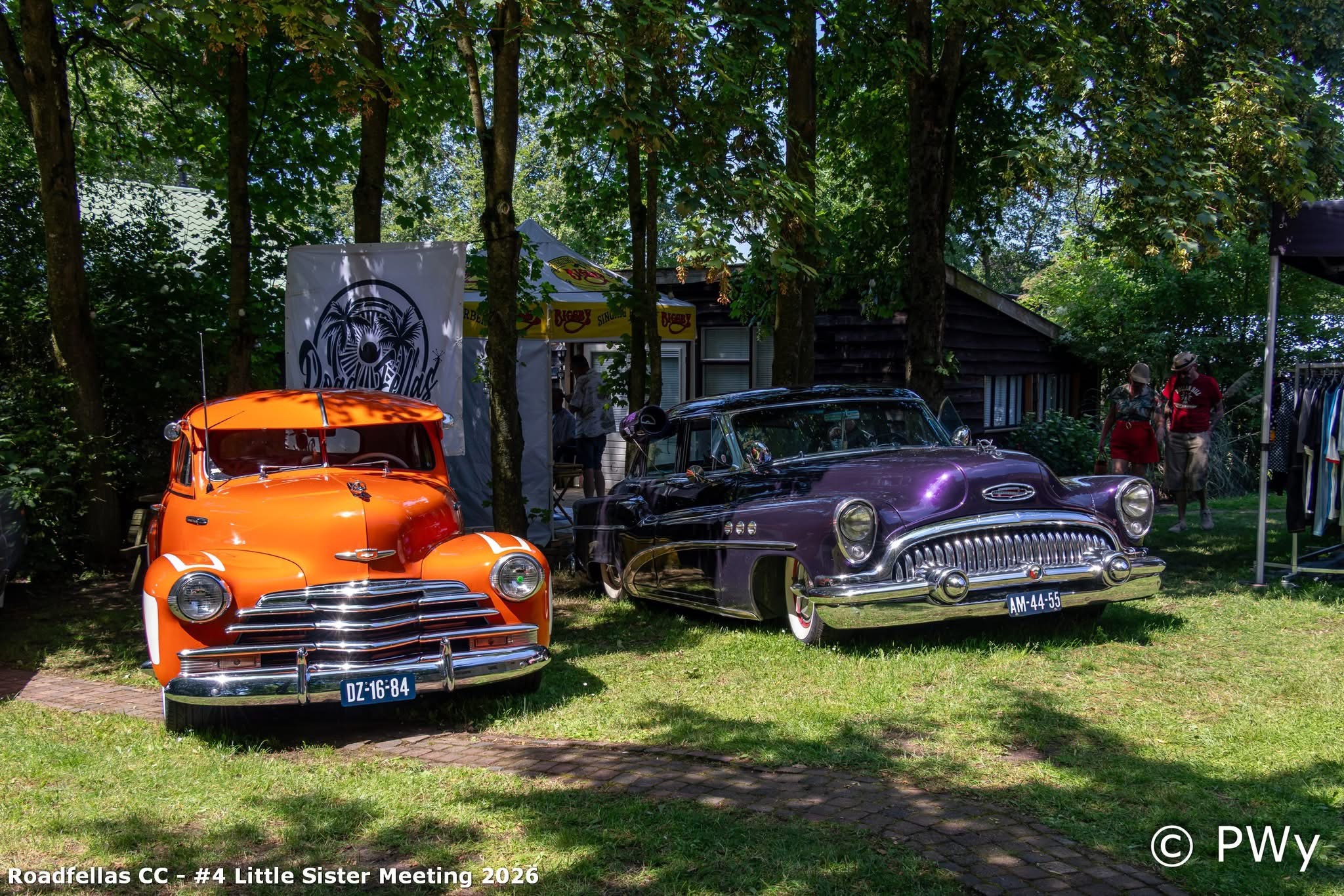 Oranje hot rod naast een paarse Buick op het gras tijdens een privé bijeenkomst