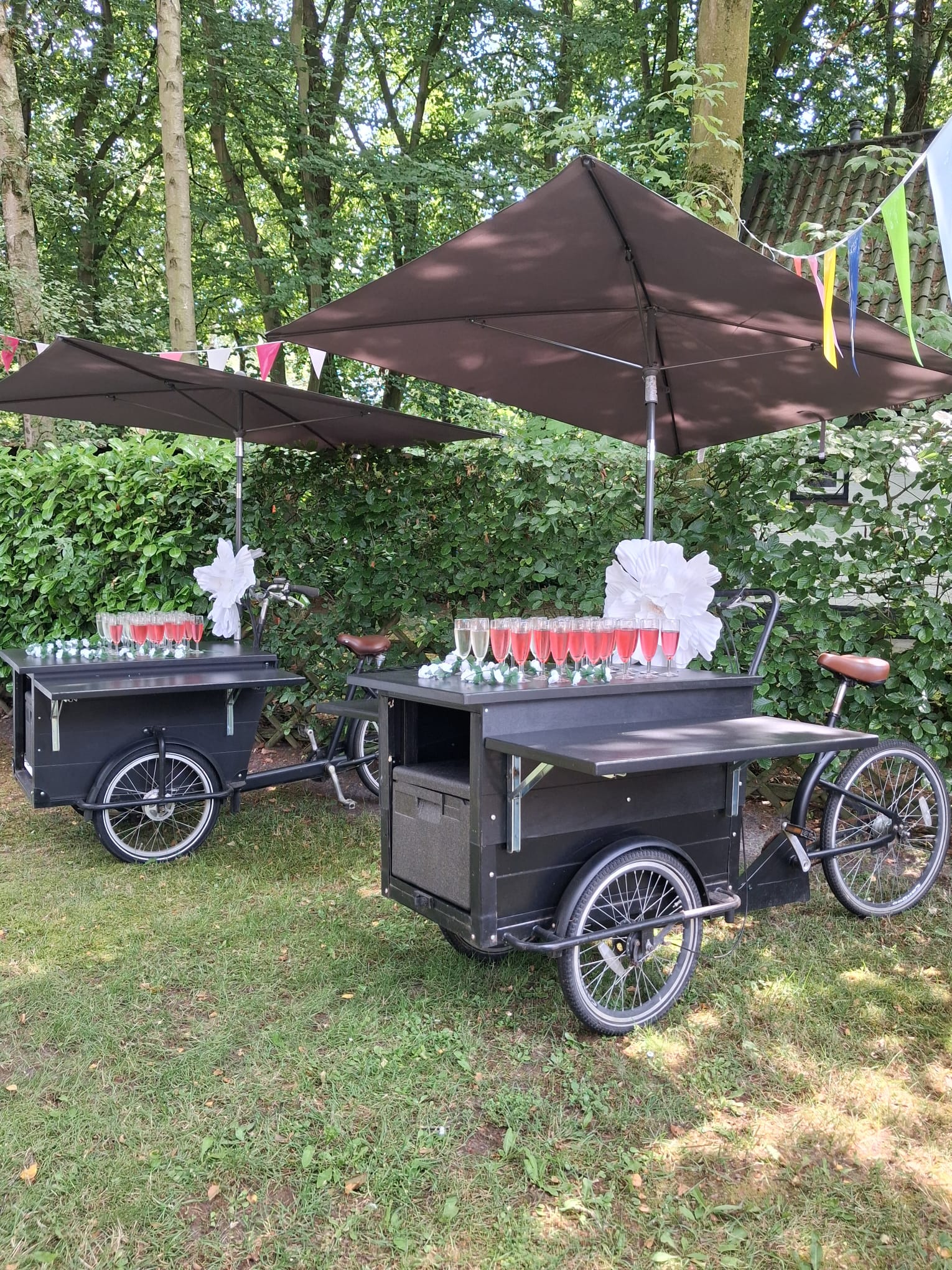 Twee zwarte bar bakfietsen met champagne en roze bubbels in glazen onder parasols