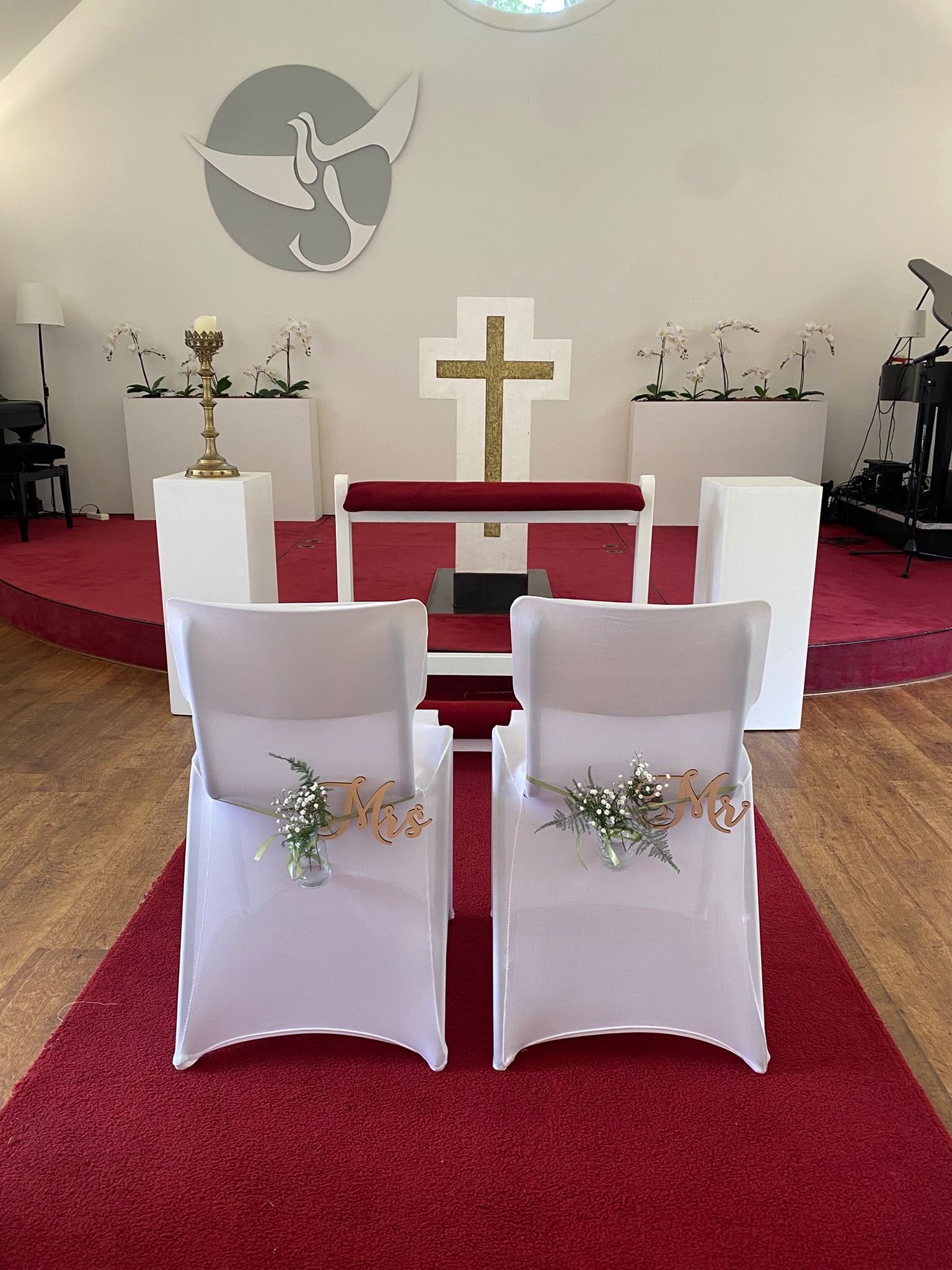 Mr & Mrs stoelen voor het altaar met kruis en witte orchideeën, duivensymbool aan de wand