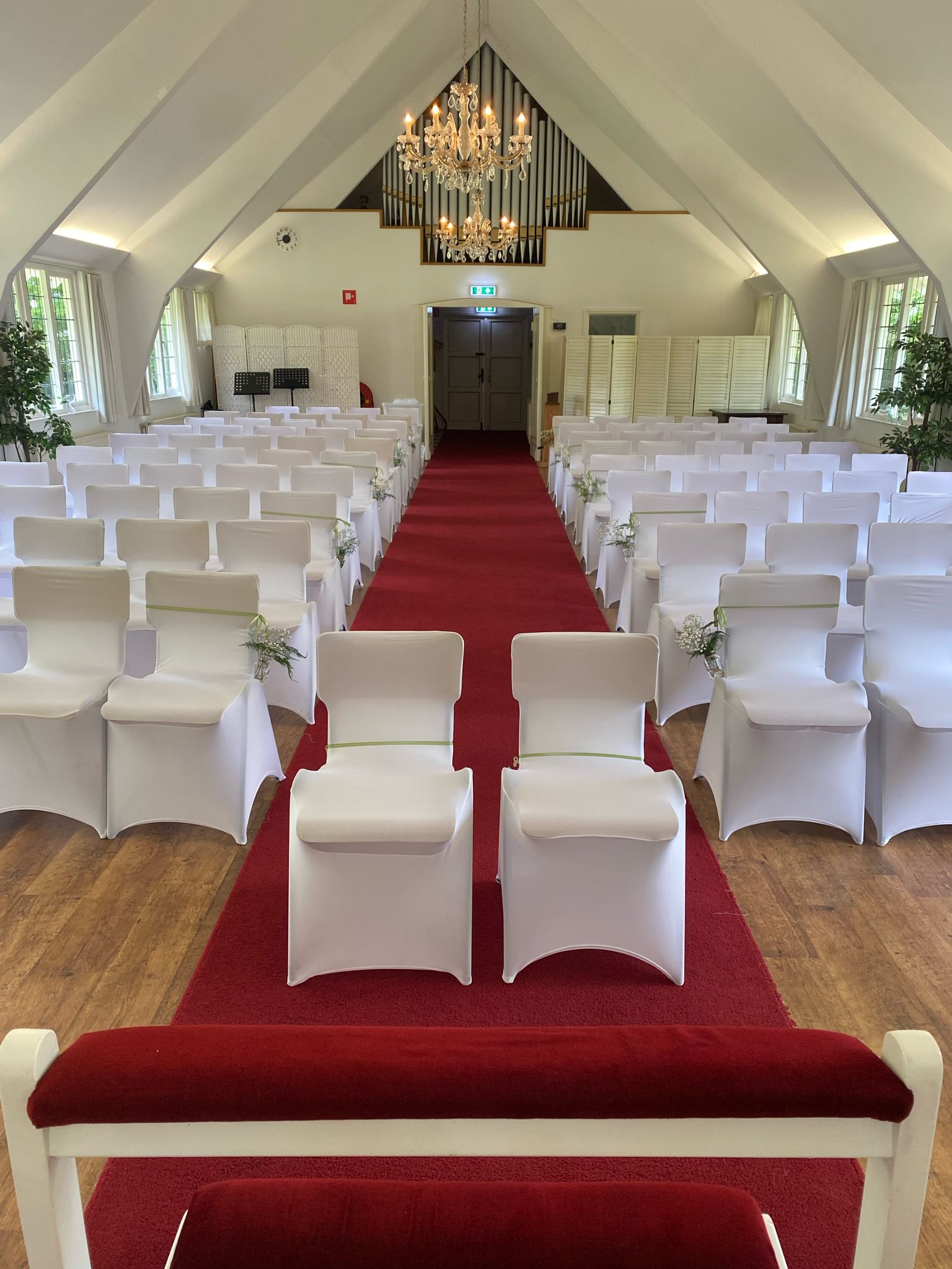 Ceremoniezaal met rijen wit gehoeste stoelen, rode middenloper, kroonluchter en orgelfront