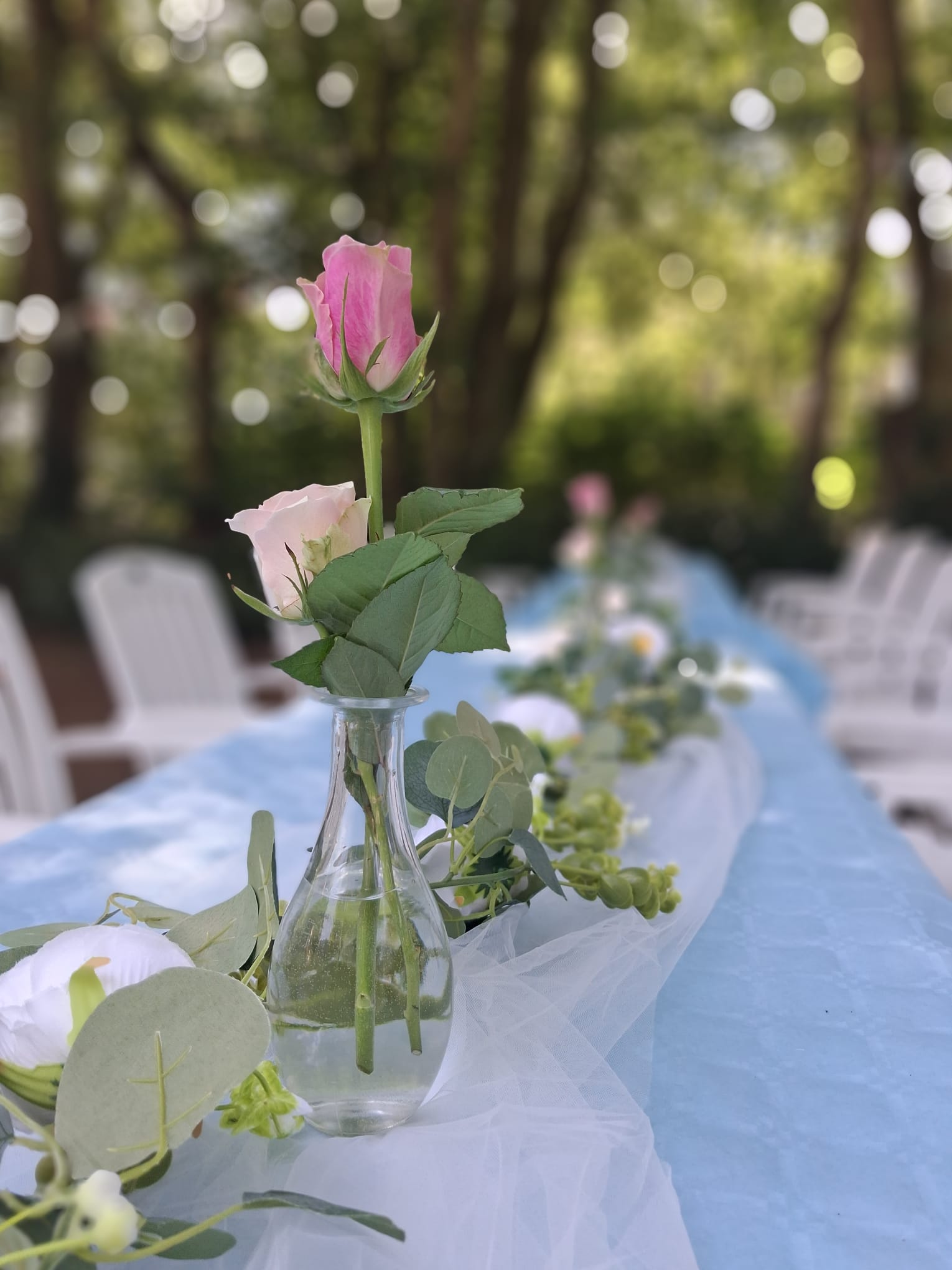 Roze rozen in een glazen vaasje op een lange gedekte tafel met lichtblauwe loper en eucalyptus in de tuin