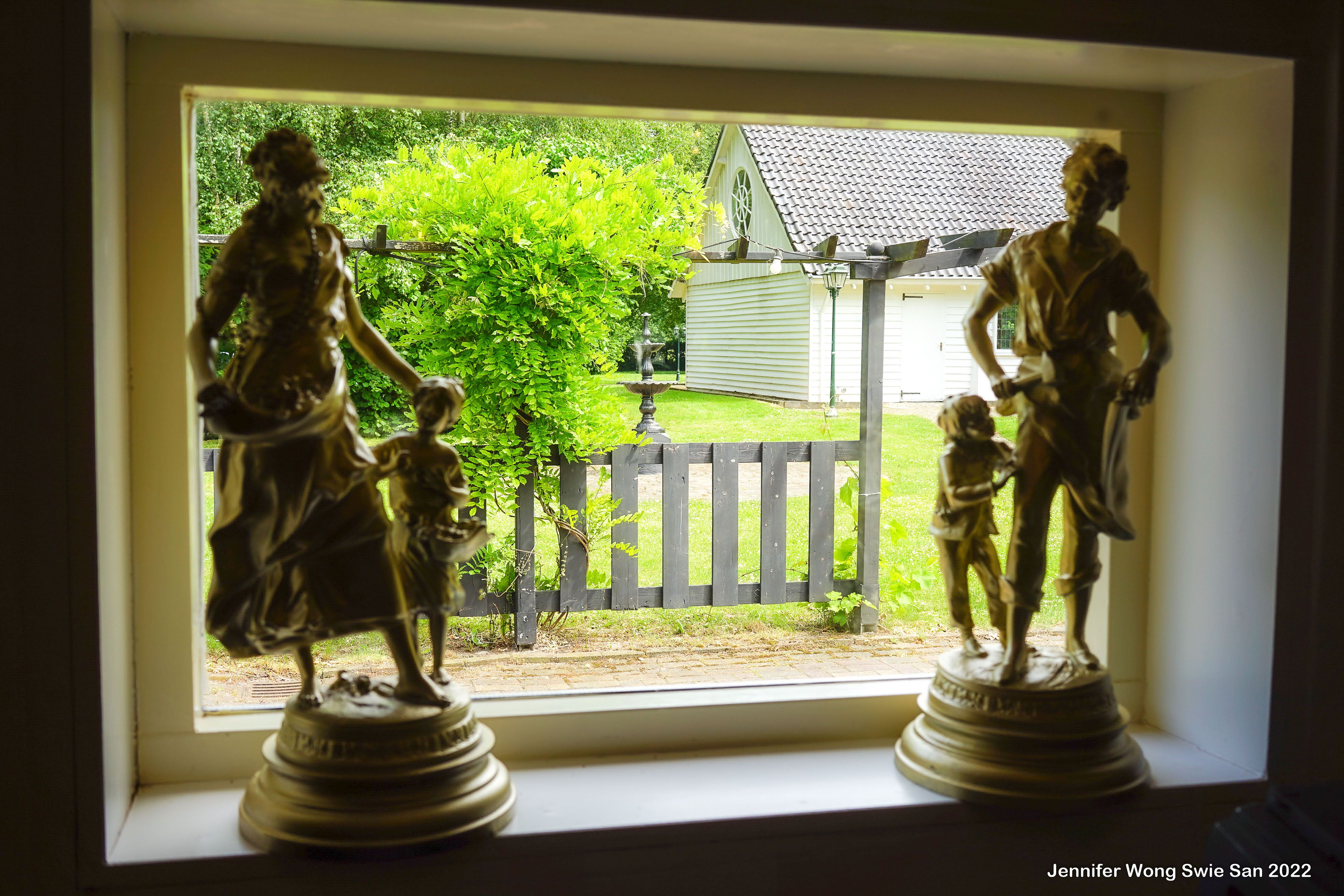 Bronzen figuurbeeldjes op een vensterbank met uitzicht op de tuin en het witte kerkje
