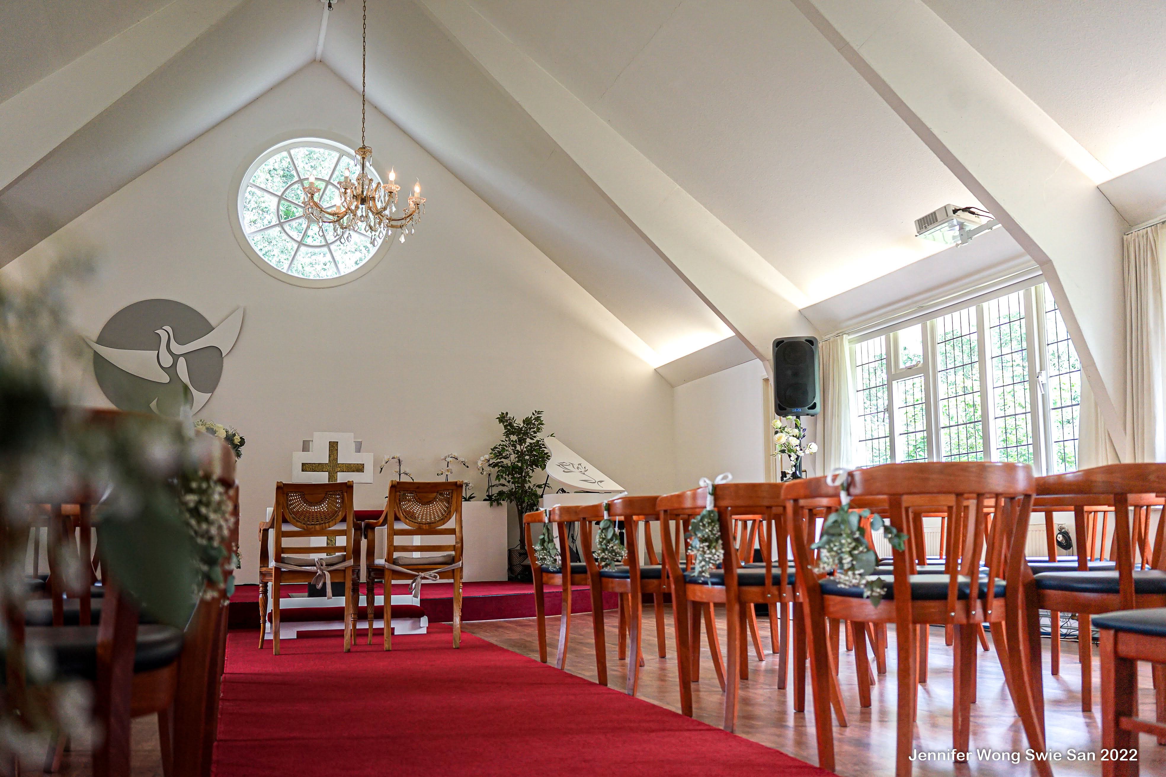 Ceremonie-zaal vanuit de zijhoek: hoog puntdak met dakbalken, rond roosvenster, kristallen kroonluchter en rijen stoelen met bloemendecoraties