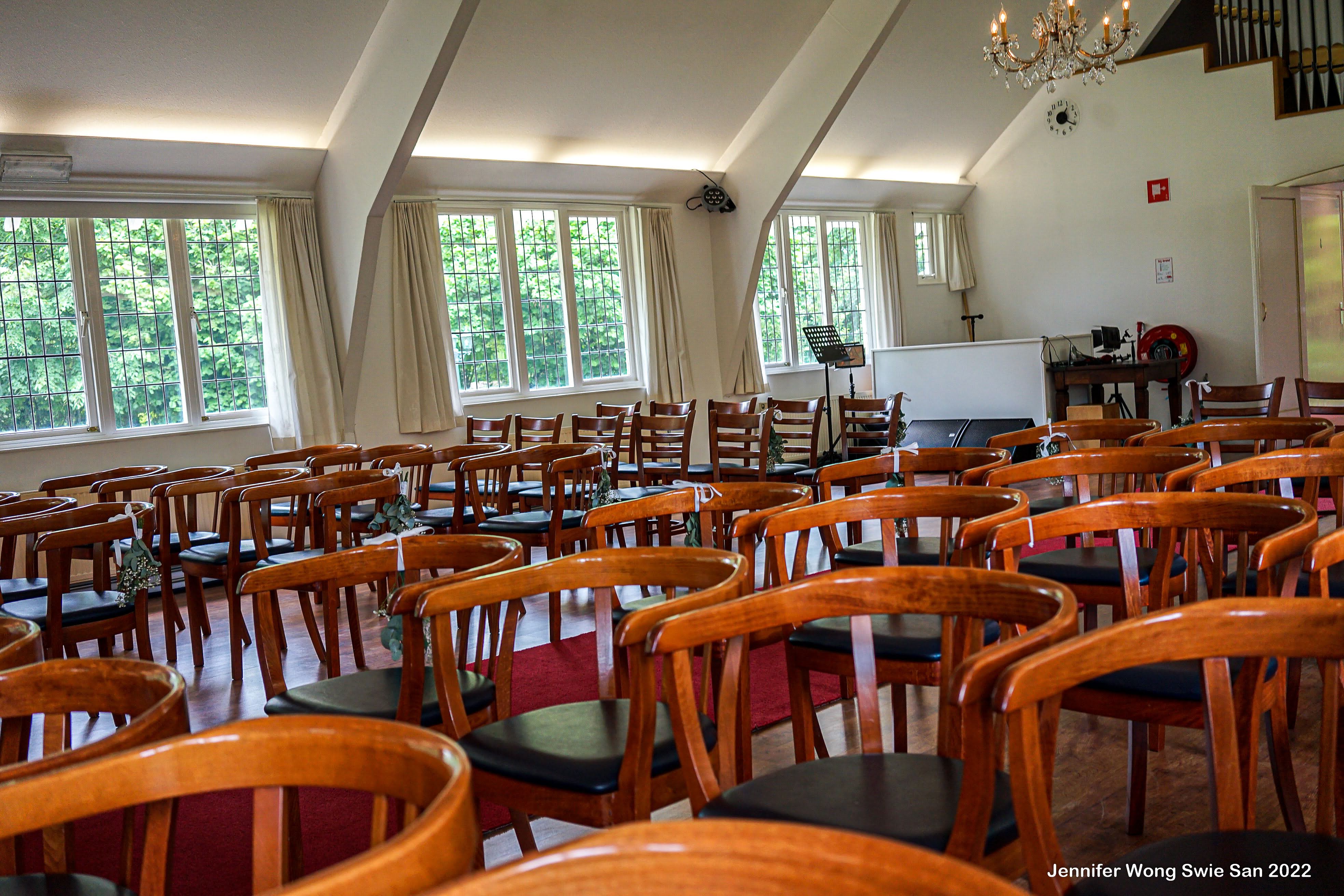 Ceremonie-zaal symmetrisch vanuit het midden richting het altaar: drie kristallen kroonluchters, groot roosvenster boven het kruis en rijen stoelen versierd met bloemen
