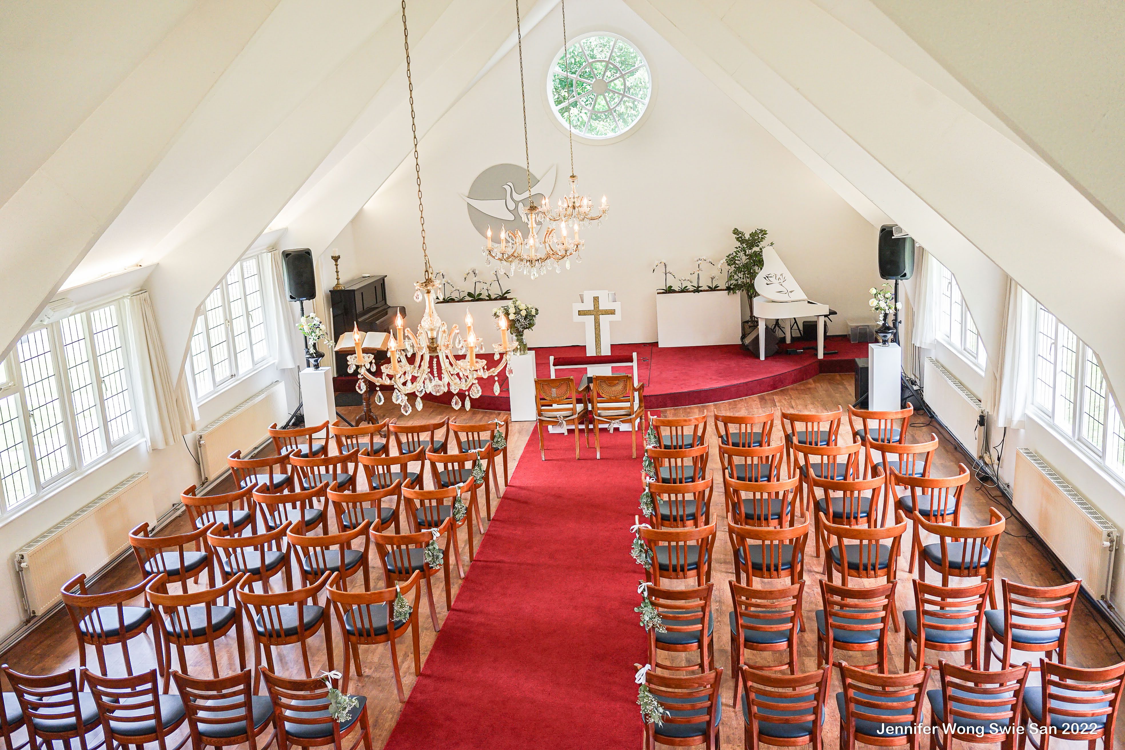 Vogelperspectief van de ceremonie-zaal bij Wit Kerkje: rode loper, alle stoelen, altaar, drie kroonluchters en piano zichtbaar